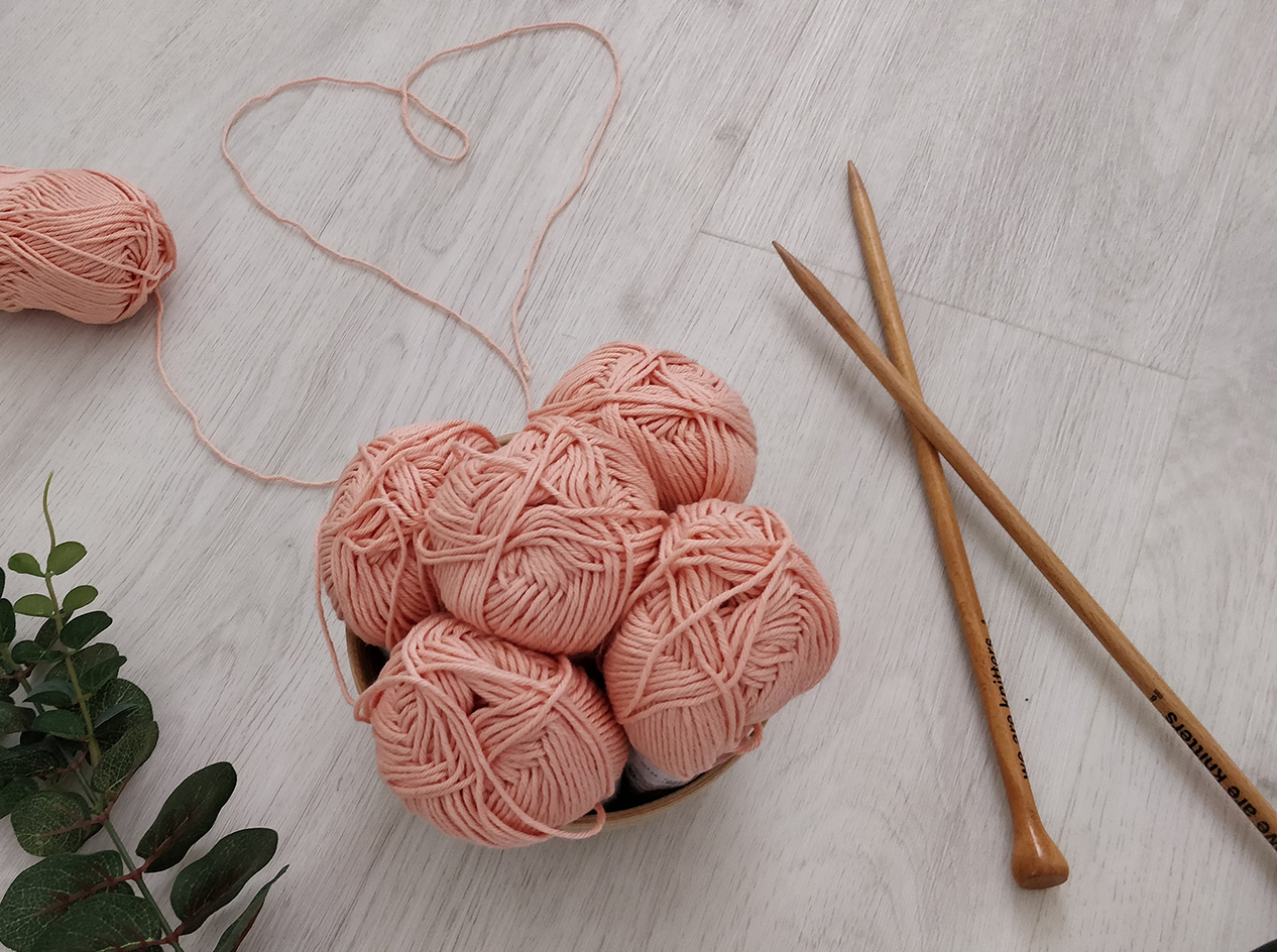 Knitting needles next to a bowl filled with pink yarn rolls.
