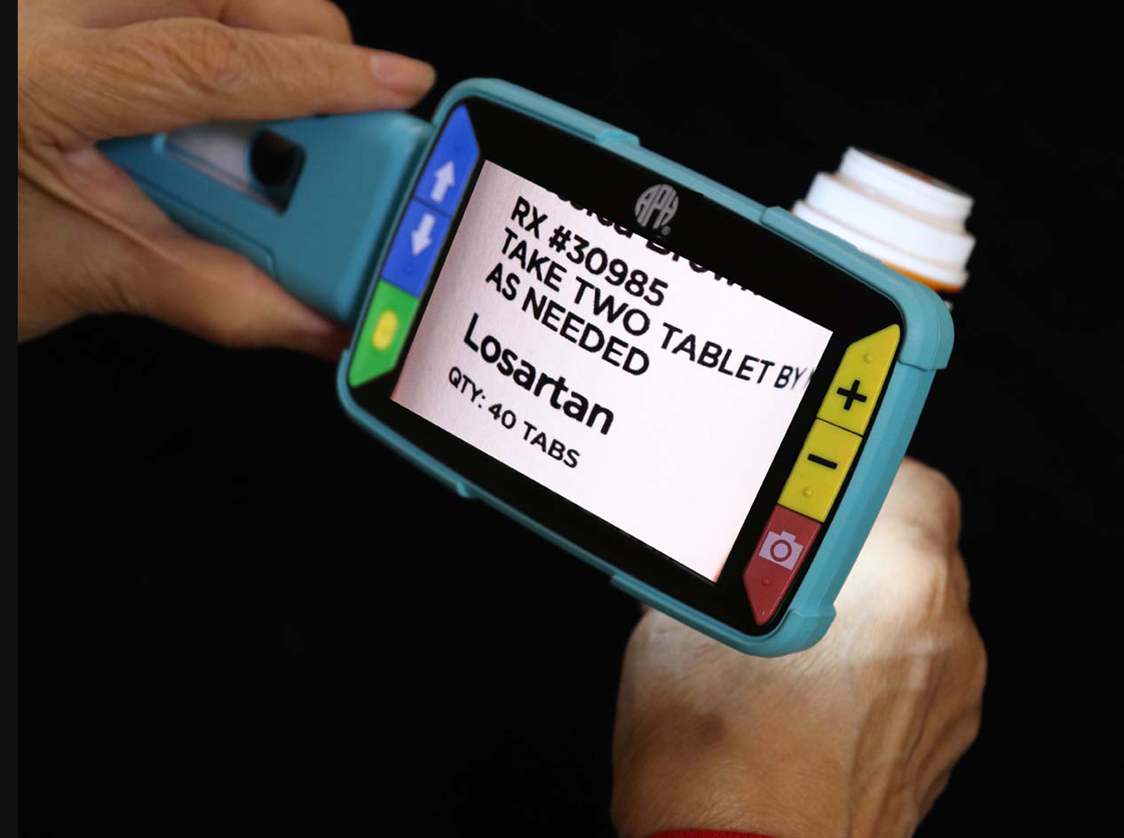 A digital handheld magnifier held up to a prescription bottle. The magnifier's screen shows an enlarged view of the prescription's information.