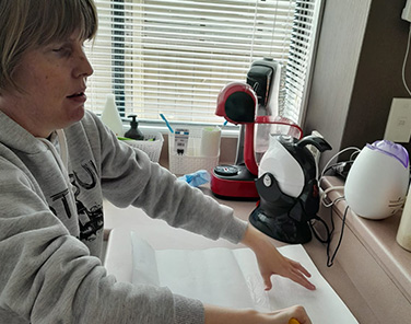 Jess Ferri, wearing a gray sweatshirt, holding a rectangle of wallpaper face down on a countertop, and using a yellow paintbrush to applying paste to the back side of the paper.