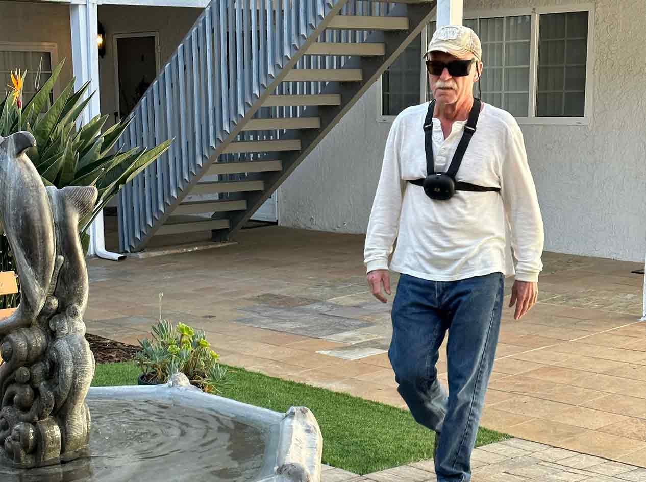 Ron Peterson, walking across a courtyard, toward a fountain. Ron is wearing an innovative navigation device called Ara, which scans the environment and provides feedback about obstacles by creating vibrations in the straps. The device is black, about the size of a fist, and the straps are fastened around the chest and over the shoulders.