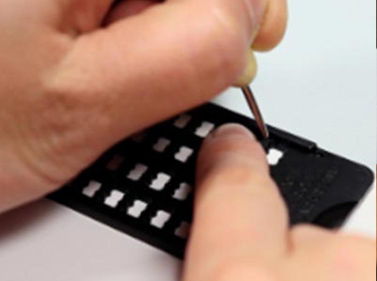 Closeup of hands using a slate and stylus to write braille. The writer is holding the stylus in the left hand and using the right index finger to guide the stylus, while punching a dot in the first cell on the right