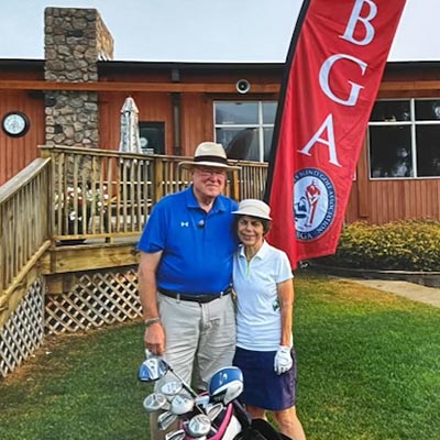 Judy Messina and her husband, Tom, pose on a lawn with Judy’s golf clubs in front of them and a bright red banner behind them. The banner has large white letters that say “USGBA,” along with the United States Blind Golf Association logo.