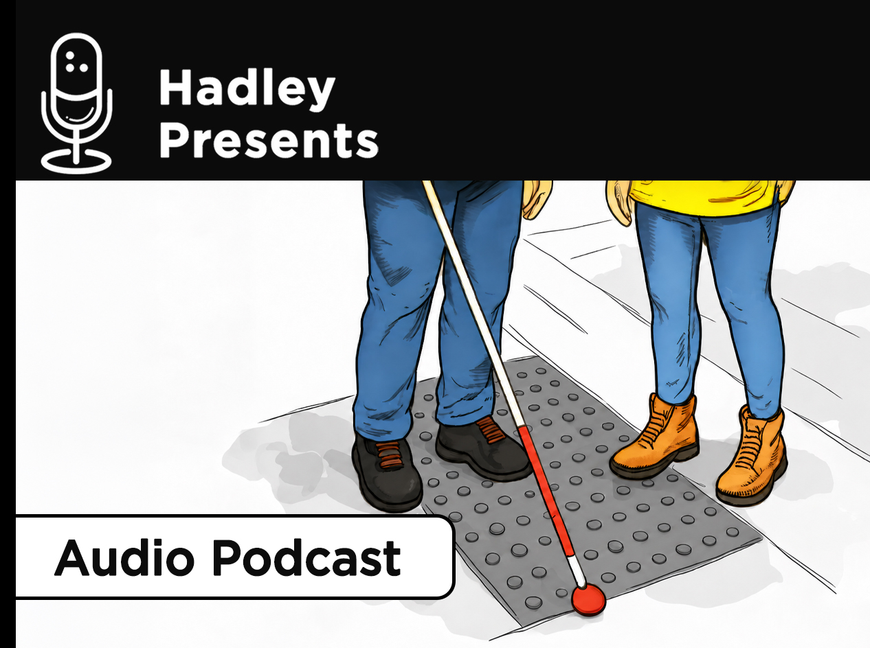 Two people stand on a sidewalk beside a dark gray tactile paving panel with raised bumps. One person, wearing blue jeans and black shoes, uses a white cane with a red tip, while the other wears blue jeans and orange boots.