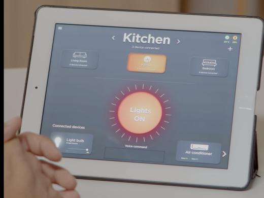 A hand reaches toward a tablet displaying a smart home control app. The screen shows the kitchen's lights turned on, with additional controls for lighting, air conditioning, and connected devices in the living room and bedroom.