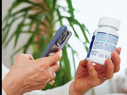 An older woman in a white sweater holds a smartphone in one hand and a white medication bottle in the other, using her phone to examine the small print on the bottle's label.