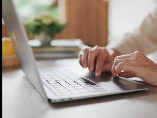 Close-up of an older adult's hands using the trackpad of a silver laptop on a wooden table.