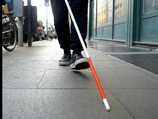 Low-angle view of a person walking along a city sidewalk using a white cane with a red tip. The person wears black pants and sneakers, with parked bicycles, cars, and buildings lining the street.