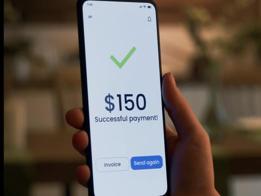 Close-up of a hand holding a smartphone displaying a green checkmark above the message "$150 Successful payment!" Two buttons labeled "Invoice" and "Send again" appear at the bottom of the screen.