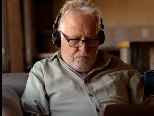 An older man with white hair, glasses, and black headphones sits on a gray couch, looking down at a laptop. He wears a light green button-down shirt, with a patterned pillow beside him and a softly lit living room in the background.