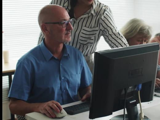 A smiling younger woman in a striped shirt assists an older man wearing glasses and a blue shirt at a desktop computer. Three other older adults work on laptops at the same table in a bright classroom with large windows.