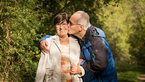 An older couple outside in a forest - the man is kissing the cheek of the woman as she smiles.