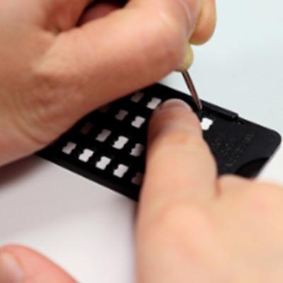 Closeup of hands using a slate and stylus to write braille. The writer is holding the stylus in the left hand and using the right index finger to guide the stylus, while punching a dot in the first cell on the right.